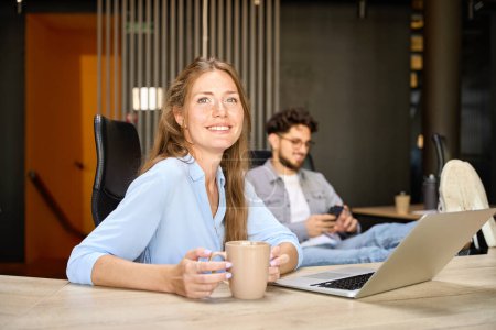 Téléchargez les photos : Smiling businesswoman looking away and drinking tea or coffee while her blurred male colleague using smartphone in coworking office. Concept of break and rest at work - en image libre de droit