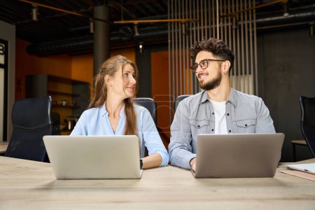 Photo for Young caucasian businessman and businesswoman looking at each other while working on laptops at desk in coworking office. Concept of teamwork - Royalty Free Image
