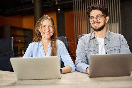 Photo for Young smiling caucasian businessman and businesswoman looking at camera while working on laptops at desk in coworking office. Concept of teamwork - Royalty Free Image