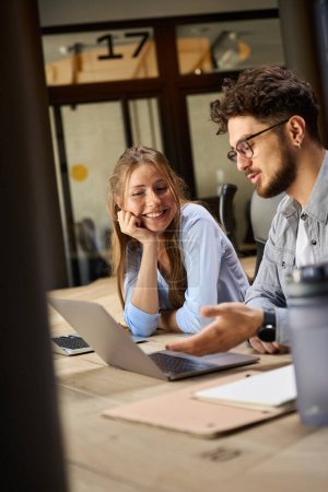 Téléchargez les photos : Young caucasian businessman showing something on laptop to smiling female colleague at desk in coworking office. Concept of teamwork - en image libre de droit