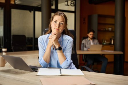 Téléchargez les photos : Young serious caucasian businesswoman looking at camera while working with laptop and notebook at desk with her blurred male colleague on background in coworking office. Modern business lifestyle - en image libre de droit