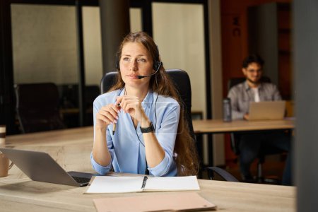 Foto de Young focused female caucasian call center operator looking away during work at desk with her blurred male colleague on background in coworking office. Concept of modern business lifestyle - Imagen libre de derechos