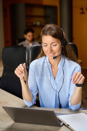 Foto de Young smiling european woman call center operator watching on laptop work at desk with her blurred male colleague on background in coworking office. Concept of modern business lifestyle - Imagen libre de derechos