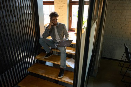 Foto de Young smiling caucasian businessman talking on mobile phone and watching on laptop on stairs in coworking office. Concept of modern business lifestyle - Imagen libre de derechos