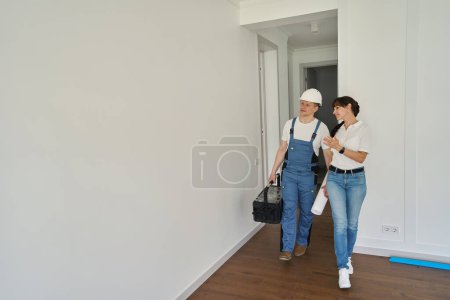 Foto de Smiling woman showing new modern comfortable townhouse for concentrated repairman with blueprint and toolbox before repair - Imagen libre de derechos
