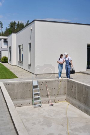 Foto de Woman and repairman talking and watching empty swimming pool before repair near new modern townhouse in warm sunny day - Imagen libre de derechos