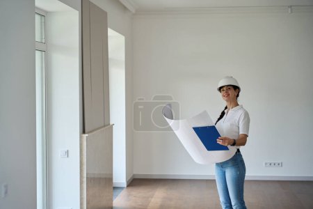 Foto de Smiling adult caucasian female repairman watching blueprint and looking at new empty modern comfortable townhouse - Imagen libre de derechos