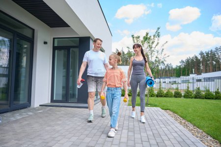 Téléchargez les photos : Smiling caucasian mother, father and daughter wearing sportswear walking on terrace near townhouse before workout in warm sunny day. Family relationship and spending time together. Healthy lifestyle - en image libre de droit