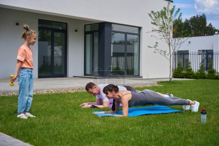 Téléchargez les photos : Little girl with dumbbells looking at her mother and father doing plank on fitness mats on green yard near townhouse in warm day. Family relationship and spending time together. Healthy lifestyle - en image libre de droit