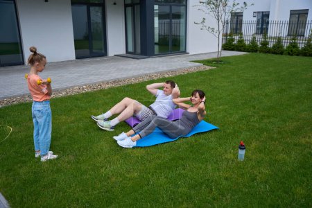 Photo for Little girl with dumbbells looking at her mom and dad doing crunching on fitness mats on green yard near townhouse in warm day. Family relationship and spending time together. Healthy lifestyle - Royalty Free Image