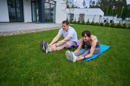 Photo for Adult caucasian couple doing stretching on fitness mats on green yard near modern townhouse in warm summer day. Family relationship and spending time together. Healthy lifestyle - Royalty Free Image