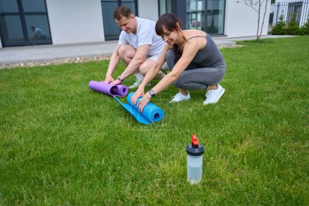Téléchargez les photos : Adult happy caucasian couple rolling up fitness mats on green yard near new modern townhouse in warm sunny day. Family relationship and spending time together. Healthy lifestyle - en image libre de droit