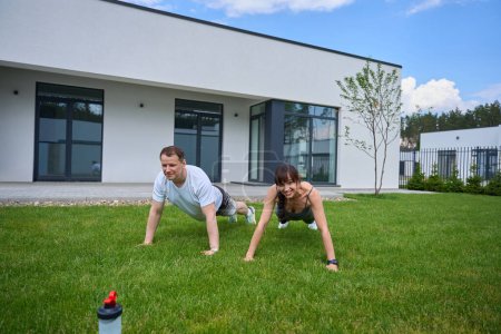 Téléchargez les photos : Adult caucasian man and smiling woman doing push ups on green grass in yard near modern townhouse in warm sunny day. Family relationship and spending time together. Healthy lifestyle - en image libre de droit