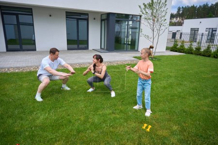 Téléchargez les photos : Little girl looking at her mother and father doing sit ups on green grass in yard near modern townhouse in warm sunny day. Family relationship and spending time together. Healthy lifestyle - en image libre de droit