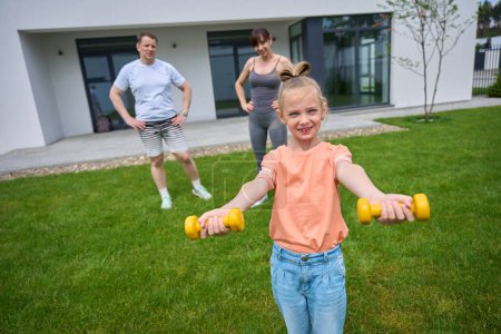Foto de Little girl exercising with dumbbells and looking at camera while her mother and father looking at daughter on green yard near townhouse in warm day. Family spending time together. Healthy lifestyle - Imagen libre de derechos