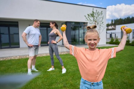Photo for Little girl exercising with dumbbells and looking at camera while her mother and father looking at each other on green yard near townhouse in warm day. Family spending time together. Healthy lifestyle - Royalty Free Image