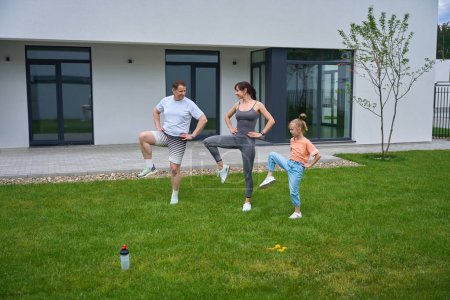 Foto de European mother, father and daughter with hands on hips doing leg exercise on green grass in yard near modern townhouse in summer day. Family relationship and spending time together. Healthy lifestyle - Imagen libre de derechos