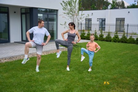 Photo for Row of caucasian mother, father and daughter with hands on hips doing leg exercise on green yard near modern townhouse in warm day. Family relationship and spending time together. Healthy lifestyle - Royalty Free Image