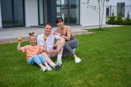 Photo for Happy caucasian mother, father and daughter sitting on green grass and looking at camera in yard near new townhouse in warm day. Family relationship and spending time together. Healthy lifestyle - Royalty Free Image