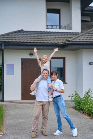 Foto de Front view of happy european mother and daughter with hands in air on fathers shoulders standing in front of modern townhouse outdoors in cloudy day. Family relationship and spending time together - Imagen libre de derechos