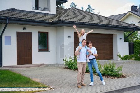 Téléchargez les photos : Pleased caucasian mother and daughter on fathers shoulders standing in front of new townhouse looking at camera outdoors in cloudy day. Family future planning, relationship and spending time together - en image libre de droit