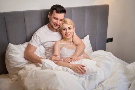 Téléchargez les photos : Young wife basks in the arms of her beloved, the couple sits on a comfortable bed - en image libre de droit