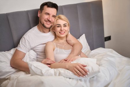 Téléchargez les photos : Young woman basks in the arms of her lover, the couple sits on a comfortable bed - en image libre de droit