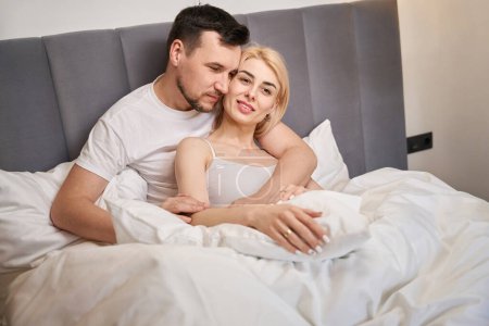 Téléchargez les photos : Young female is basking in the arms of her lover, the couple is seated on a comfortable bed - en image libre de droit