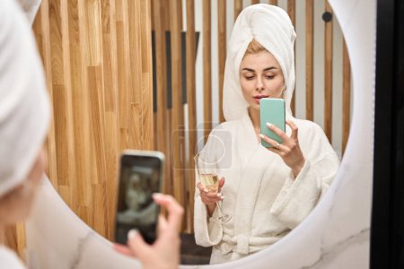 Téléchargez les photos : Blonde woman in the bathroom takes a selfie in front of the mirror, she is in a fluffy bathrobe - en image libre de droit