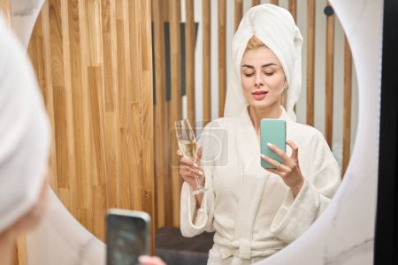 Téléchargez les photos : Woman in the bathroom takes a selfie in front of the mirror, she is in a fluffy bathrobe - en image libre de droit