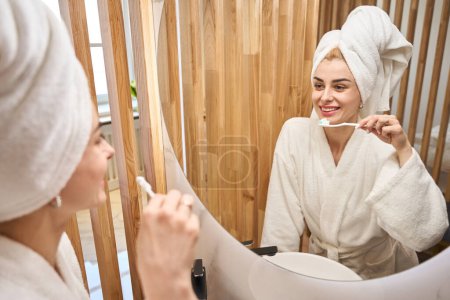 Foto de Woman brushes her teeth in front of a mirror, she is in a fluffy bathrobe - Imagen libre de derechos