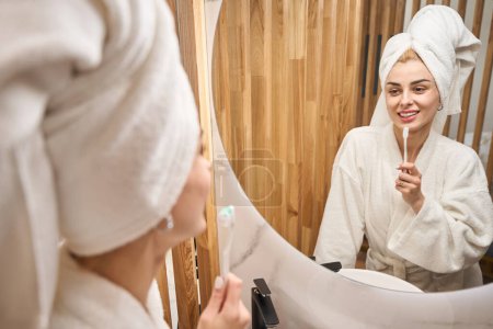 Téléchargez les photos : Female with a towel turban brushes her teeth in front of a mirror, she is in a fluffy bathrobe - en image libre de droit