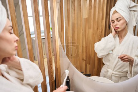 Photo for Female with her eyes closed stands with a toothbrush in front of a mirror, she is in a fluffy bathrobe - Royalty Free Image