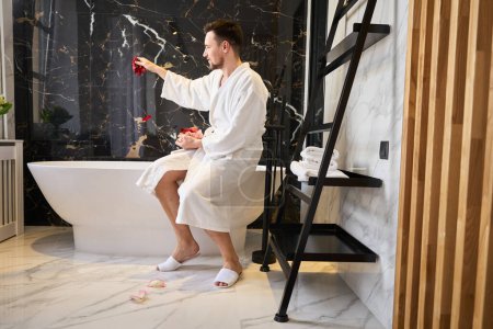 Téléchargez les photos : Male in a fluffy bathrobe sits on the edge of the bathtub, he prepares a bath with rose petals - en image libre de droit