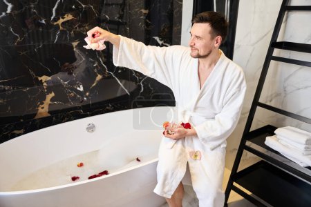Téléchargez les photos : Smiling male in a fluffy bathrobe sits on the edge of the bathtub, he prepares a bath with rose petals - en image libre de droit