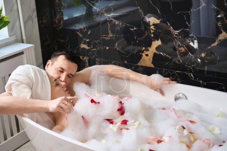 Photo for Newlyweds soak in a foam bath with rose petals, a man gently hugs and kisses a woman - Royalty Free Image