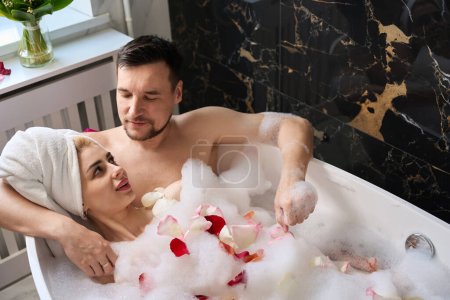Foto de Newlyweds enjoy a bubble bath with rose petals, a man gently hugs his wife - Imagen libre de derechos