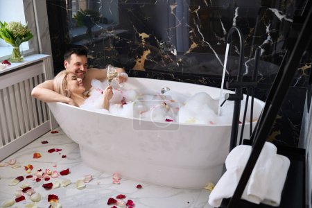 Téléchargez les photos : Newlyweds enjoy champagne in a foam bath with rose petals, a man tenderly hugs a woman - en image libre de droit