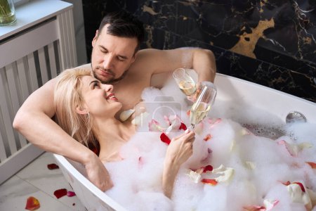 Téléchargez les photos : Joyful newlyweds enjoying champagne in a bathtub with rose petals, a man tenderly hugs a woman - en image libre de droit