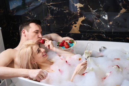 Téléchargez les photos : Female and a male feast on strawberries and champagne in a foam bath, a man gently hugs a woman - en image libre de droit