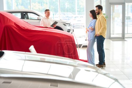 Foto de Car dealership consultant presents a car under a red cover, a buyer and his wife are waiting for a surprise - Imagen libre de derechos