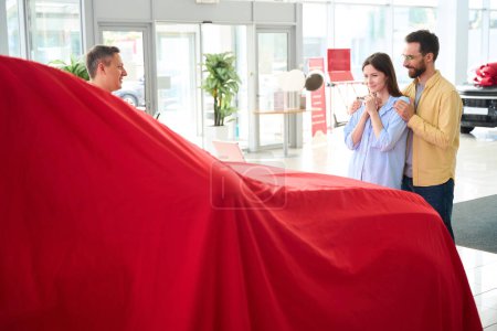 Foto de Customer service manager presents the car under a red cover, the spouses are waiting for a surprise - Imagen libre de derechos