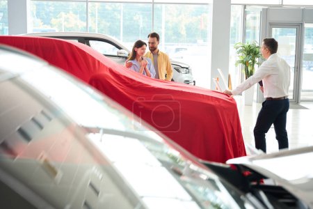 Foto de Manager of car dealership presents the car under red cover, the buyer and his wife are waiting for a surprise - Imagen libre de derechos