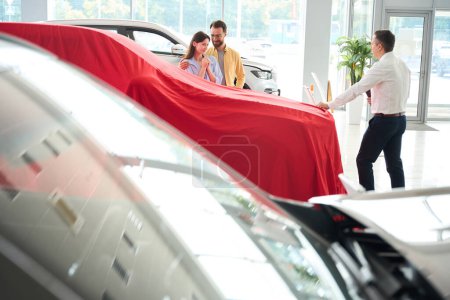 Téléchargez les photos : Car dealership employee presents a car with a red cover, a buyer and his wife are waiting for a surprise - en image libre de droit