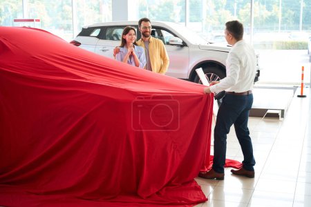 Photo for Car dealership employee removes a gift cover from a car, the buyer has prepared a surprise for his wife - Royalty Free Image