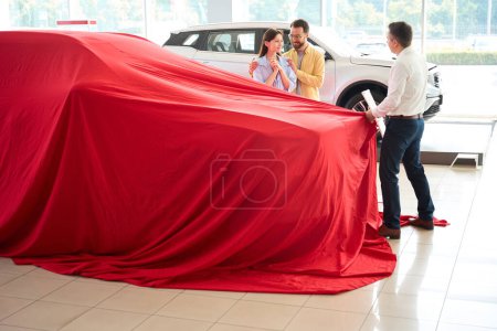 Foto de Car dealership employee removes a red cover from a car, the buyer has prepared a surprise for his wife - Imagen libre de derechos