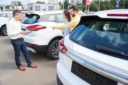 Photo for People inspect a car in the yard of a car dealership, a consultant helps them - Royalty Free Image