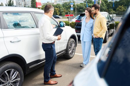 Photo for Married couple inspects a car in the yard of a car dealership, a consultant helps them - Royalty Free Image