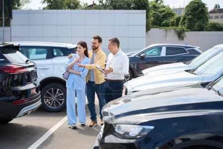 Téléchargez les photos : Husband and wife choose a car in the yard of a car dealership, a consultant helps them - en image libre de droit
