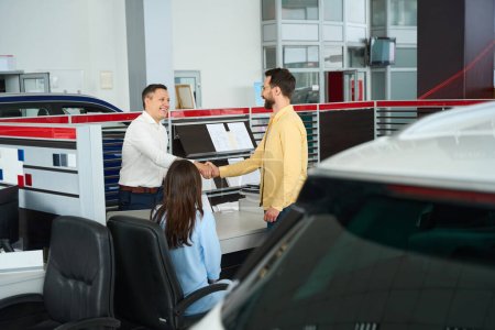 Foto de Consultant greets a client in the office area of a car dealership, the buyers wife is sitting nearby - Imagen libre de derechos